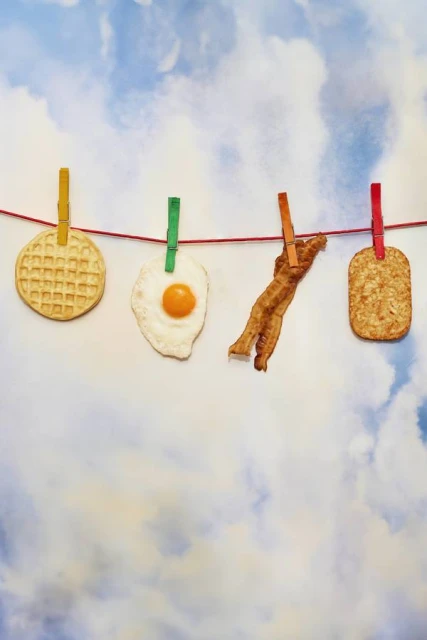 Overhead shot of a waffle, fried egg, bacon, and hash brown hung on a clothesline with colorful clothespins against a cloudy sky background.