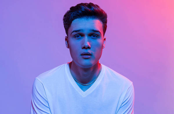 Portrait of a young man with short brown hair and a white t-shirt against a gradient pink and blue background in a photo studio.