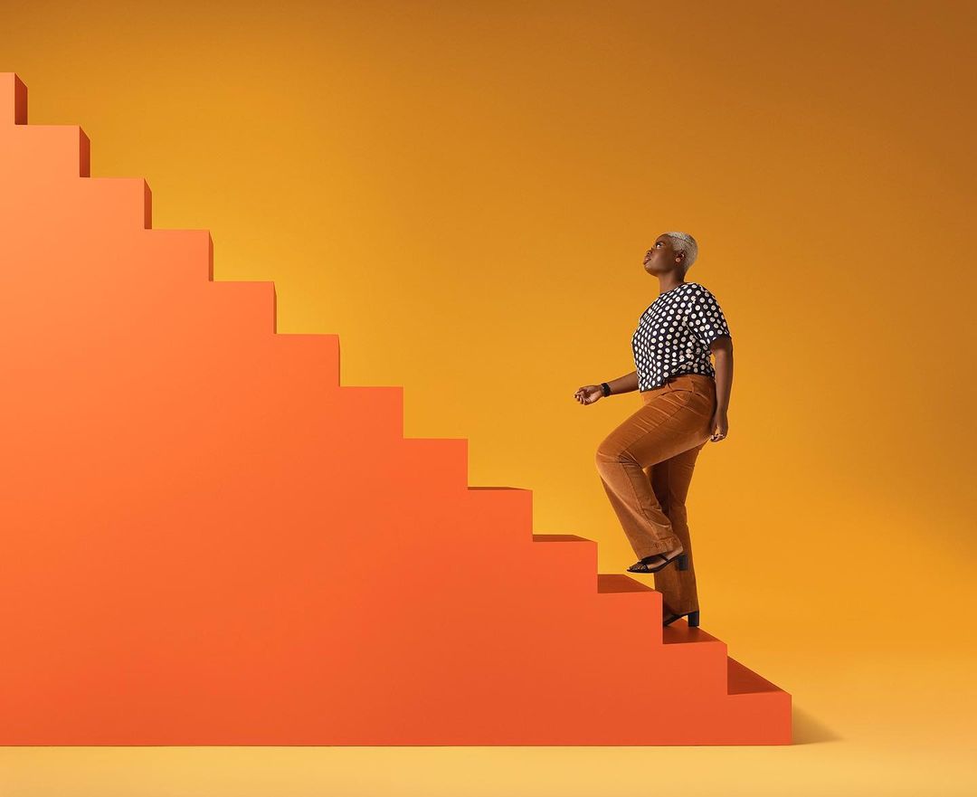 An African-American woman ascends bright orange stairs against a yellow background in a studio shot, promoting health and lifestyle.