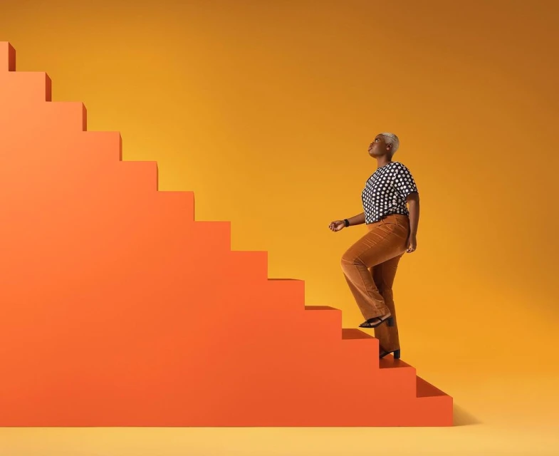 An African-American woman ascends bright orange stairs against a yellow background in a studio shot, promoting health and lifestyle.