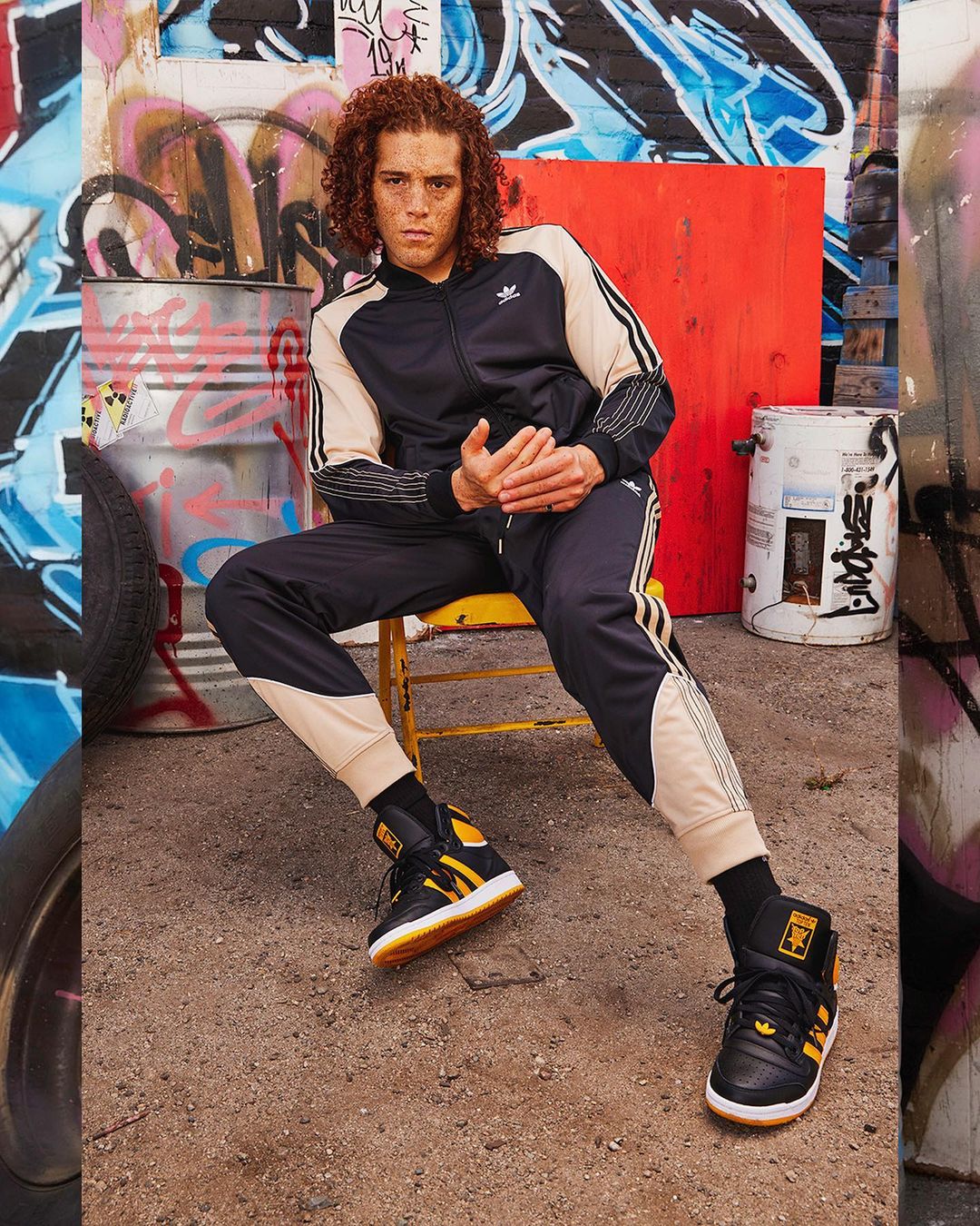 Man with curly hair in black and beige tracksuit and sneakers sitting on a yellow chair, graffiti and metal cans background.