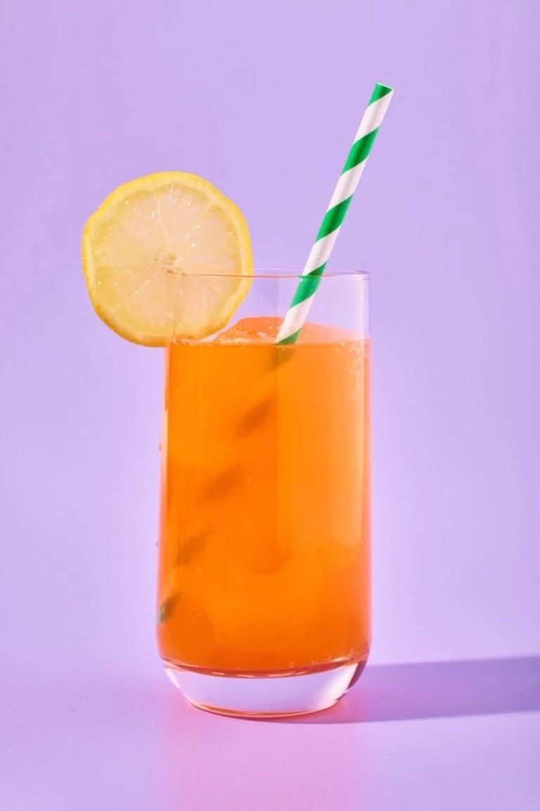 Refreshing orange beverage in a glass with lemon slice and striped straw, set against a light purple background in a studio shot.