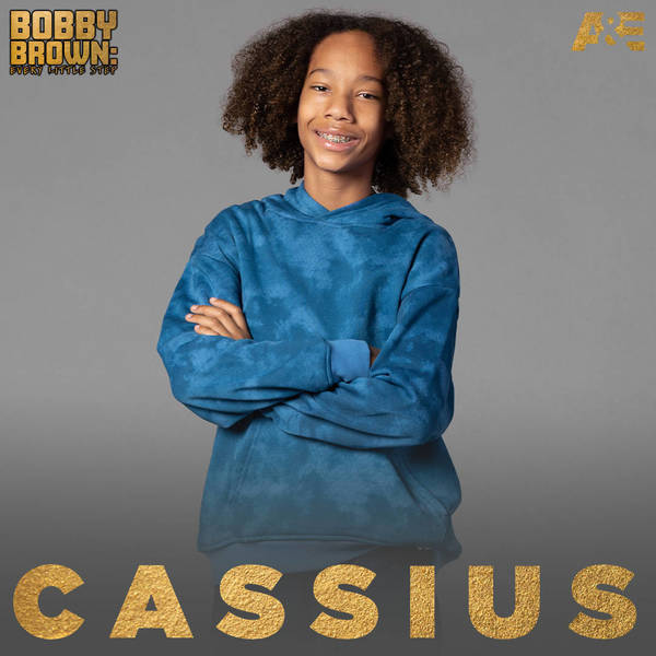 Portrait of a smiling young boy with curly hair wearing a blue hoodie with arms crossed against a gray background with gold text.