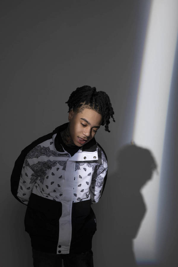 Portrait of a Black man with dreadlocks wearing a patterned black and white jacket against a grey wall with a vertical beam of light