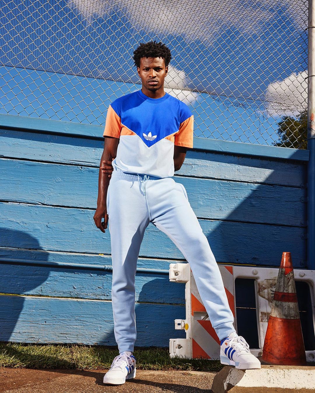 Full-length portrait of a young Black man in a blue, peach, and white color-blocked t-shirt and light blue sweatpants and sneakers.