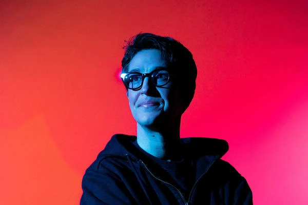 Adult woman with short hair and glasses smiles in a close-up portrait against a red and blue gradient background in an indoor setting.
