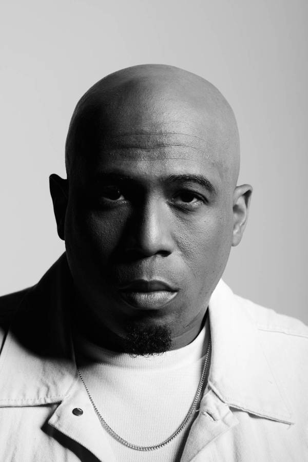 Black and white portrait of a bald adult male with a goatee, wearing a white shirt and chain necklace against a neutral background.