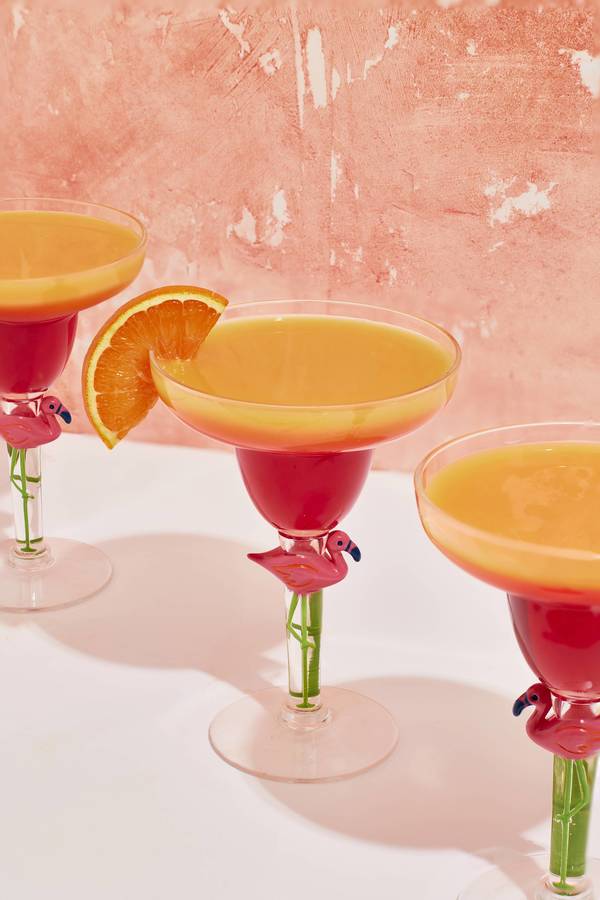 Three cocktail glasses with flamingo stems hold orange drinks, one garnished with orange slice, against a pink textured background.