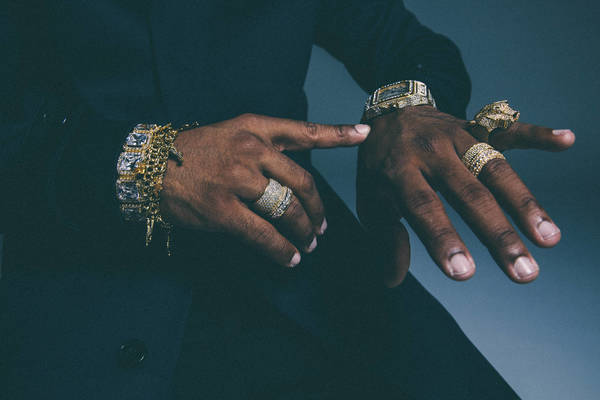 Close-up shot of an adult's dark-skinned hands displaying a gold watch and multiple gold rings and bracelets, indicating wealth and style.