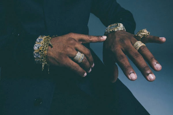 Close-up shot of an adult's dark-skinned hands displaying a gold watch and multiple gold rings and bracelets, indicating wealth and style.