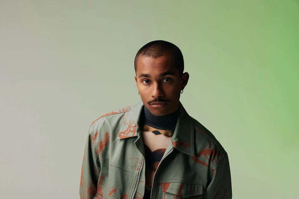 Portrait of a young adult male in a patterned jacket and turtleneck against a graduated green background, looking directly at the camera.