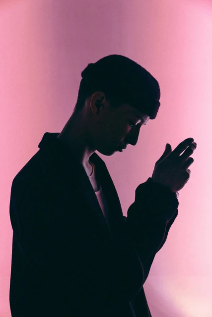 Silhouette of a male adult with a neutral expression wearing a beanie and coat, lit with pink lighting against a pink background.