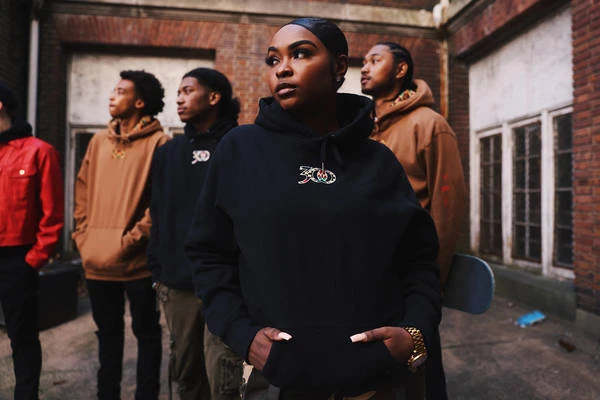 Group of young adults wearing hoodies standing outdoors near a brick building with windows.