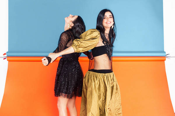 Two smiling women in dresses pose back-to-back against a background of bright orange and blue seamless paper, studio lighting.