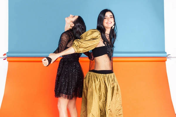 Two smiling women in dresses pose back-to-back against a background of bright orange and blue seamless paper, studio lighting.