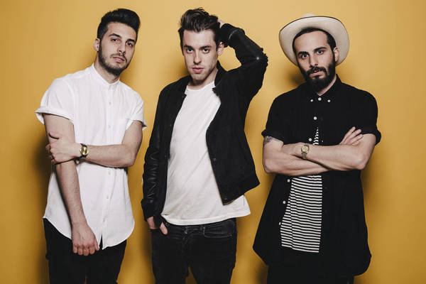 Three young adult men pose in front of a yellow background. Two are wearing white shirts and one is wearing a hat.