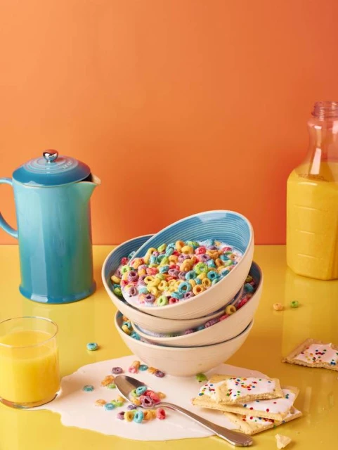 Stacked bowls of colorful cereal with milk on a yellow surface.  Orange juice and Pop-Tarts are also visible in the food still life.