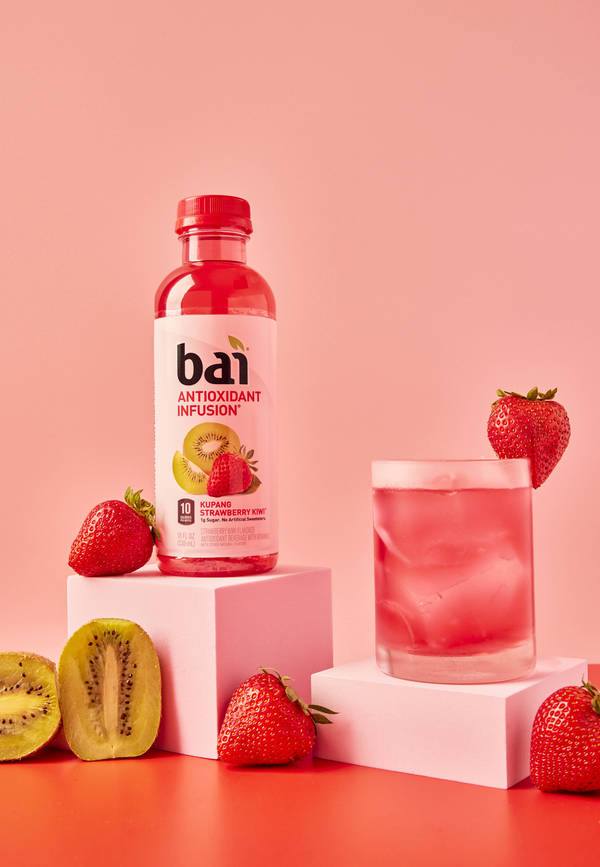 Product shot of Bai antioxidant drink and glass with pink liquid and ice, surrounded by strawberries and cut kiwi on a pink background.