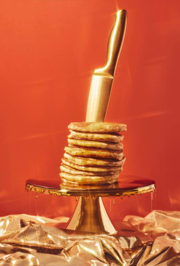 Still life shot of stacked pancakes on a golden cake stand, topped with golden syrup and skewered with a golden knife, against red background.