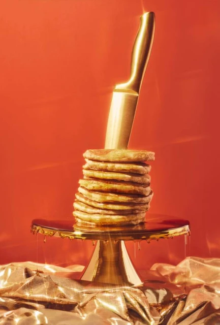 Still life shot of stacked pancakes on a golden cake stand, topped with golden syrup and skewered with a golden knife, against red background.