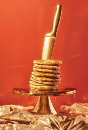 Still life shot of stacked pancakes on a golden cake stand, topped with golden syrup and skewered with a golden knife, against red background.