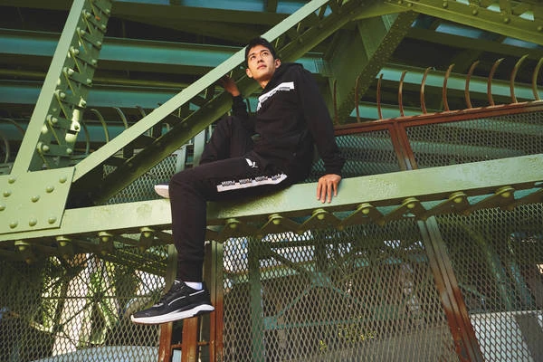 Young man in Puma tracksuit and sneakers sitting on green steel bridge, looking up; urban environment with metal and structural lines.