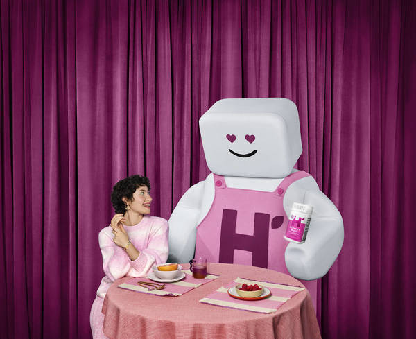 Woman sitting at a table with cartoon character, plates of food, pink tablecloth, and pink curtains in the background.