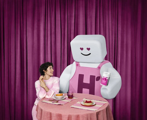 Woman sitting at a table with cartoon character, plates of food, pink tablecloth, and pink curtains in the background. 