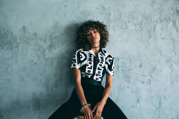 Young woman with curly hair posing against a mottled grey wall wearing a patterned shirt, black pants, and gold jewelry.