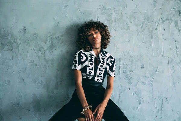 Young woman with curly hair posing against a mottled grey wall wearing a patterned shirt, black pants, and gold jewelry.