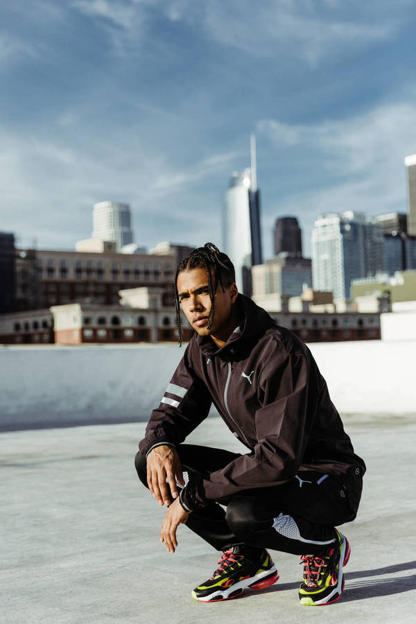 Athletic man in black Puma tracksuit and multicolored sneakers crouches against the backdrop of a city skyline with blue sky and clouds.