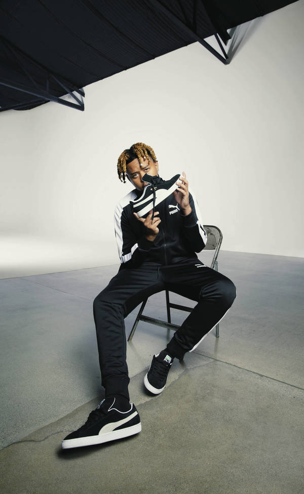 Young man in black tracksuit sitting and holding a sneaker in a photo studio with black and white colors.