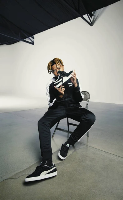 Young man in black tracksuit sitting and holding a sneaker in a photo studio with black and white colors.