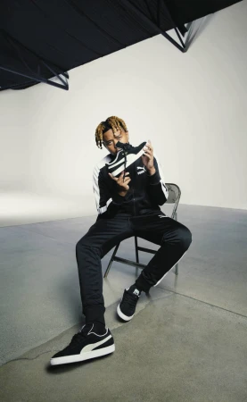 Young man in black tracksuit sitting and holding a sneaker in a photo studio with black and white colors.
