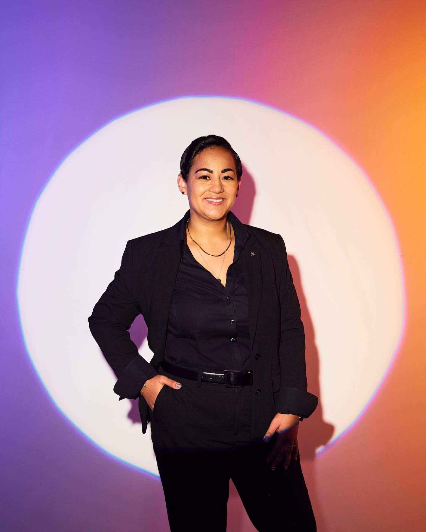 Woman in black suit poses in front of a spotlight with a purple and orange gradient background in a portrait-style studio shot.
