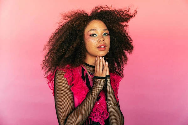 Portrait of a young woman with curly hair against a pink backdrop, wearing a pink vest and black fishnet sleeves, beauty and fashion.