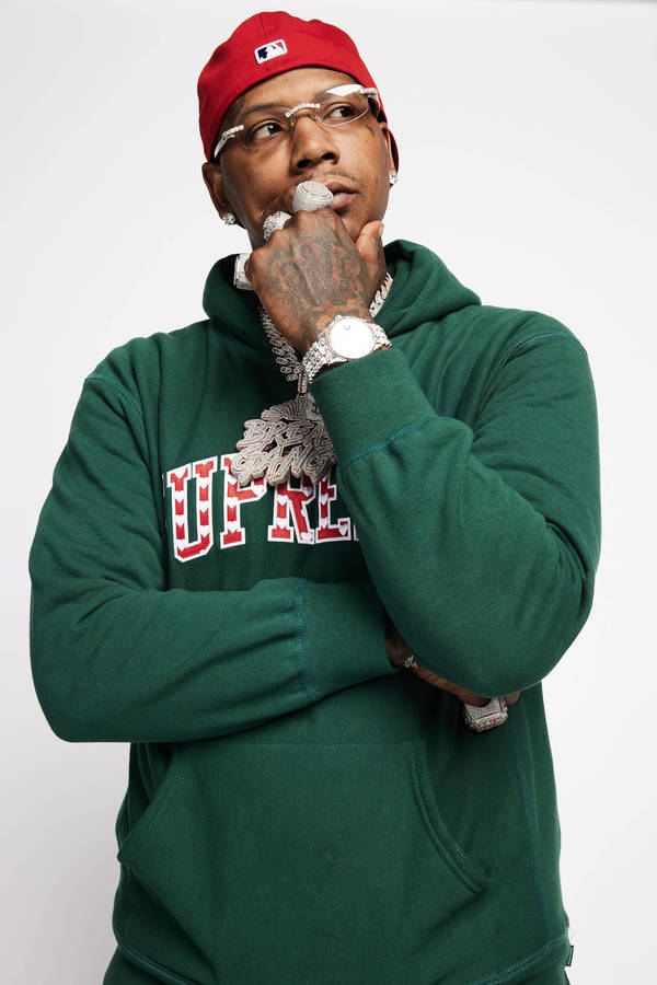 Portrait of a man with glasses and a red cap, wearing a green hoodie and a diamond chain, posing against a plain white background.