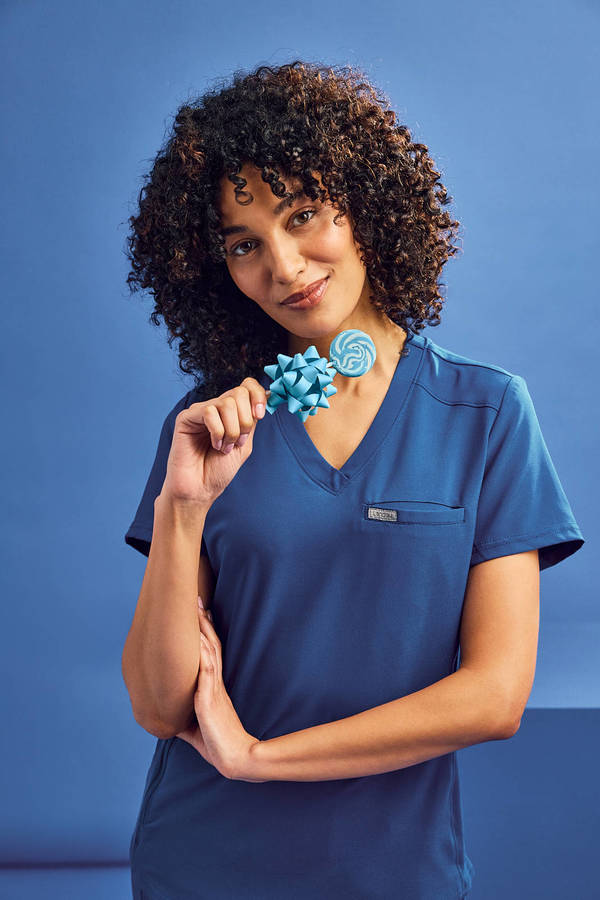 A woman in a blue scrub top smiles, holding a blue bow and lollipop against a matching blue backdrop, portrait framing, eye-level.