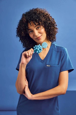 A woman in a blue scrub top smiles, holding a blue bow and lollipop against a matching blue backdrop, portrait framing, eye-level.