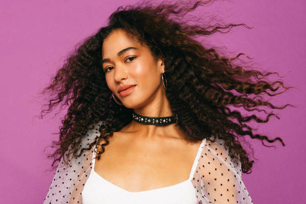 Portrait of a smiling woman with curly brown hair, wearing a white top and black choker against a purple backdrop in a photo studio.