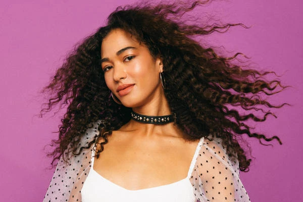 Portrait of a smiling woman with curly brown hair, wearing a white top and black choker against a purple backdrop in a photo studio.