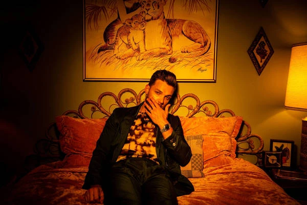 A man in a patterned shirt and black jacket sits on a bed under a framed painting of lions in a dimly lit room.