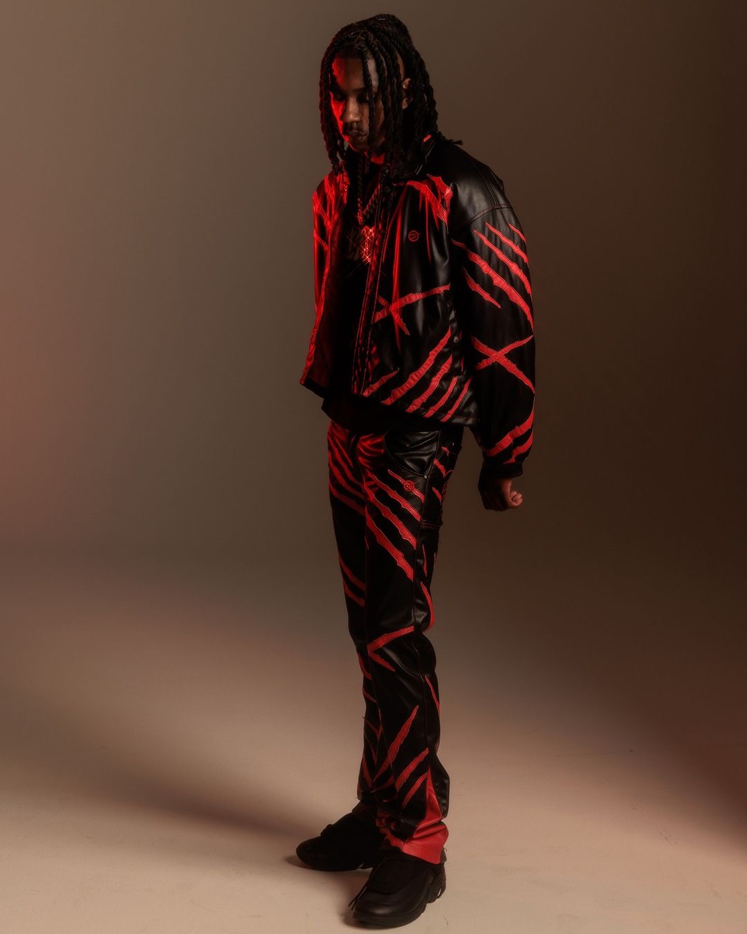 A stylish adult male poses in a black leather jacket and pants with red stripes, illuminated by red lighting against a neutral backdrop.