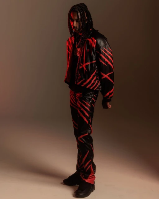 A stylish adult male poses in a black leather jacket and pants with red stripes, illuminated by red lighting against a neutral backdrop.