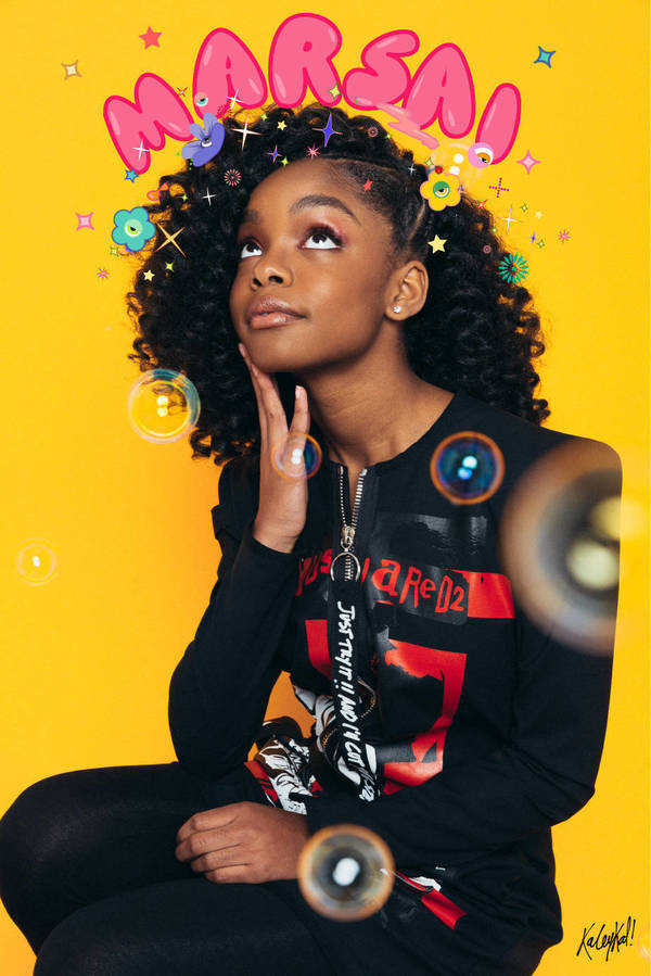 Young Black girl with afro sits in front of a yellow backdrop, wearing a black sweatshirt with graphics, accessorized with floating bubble effects.