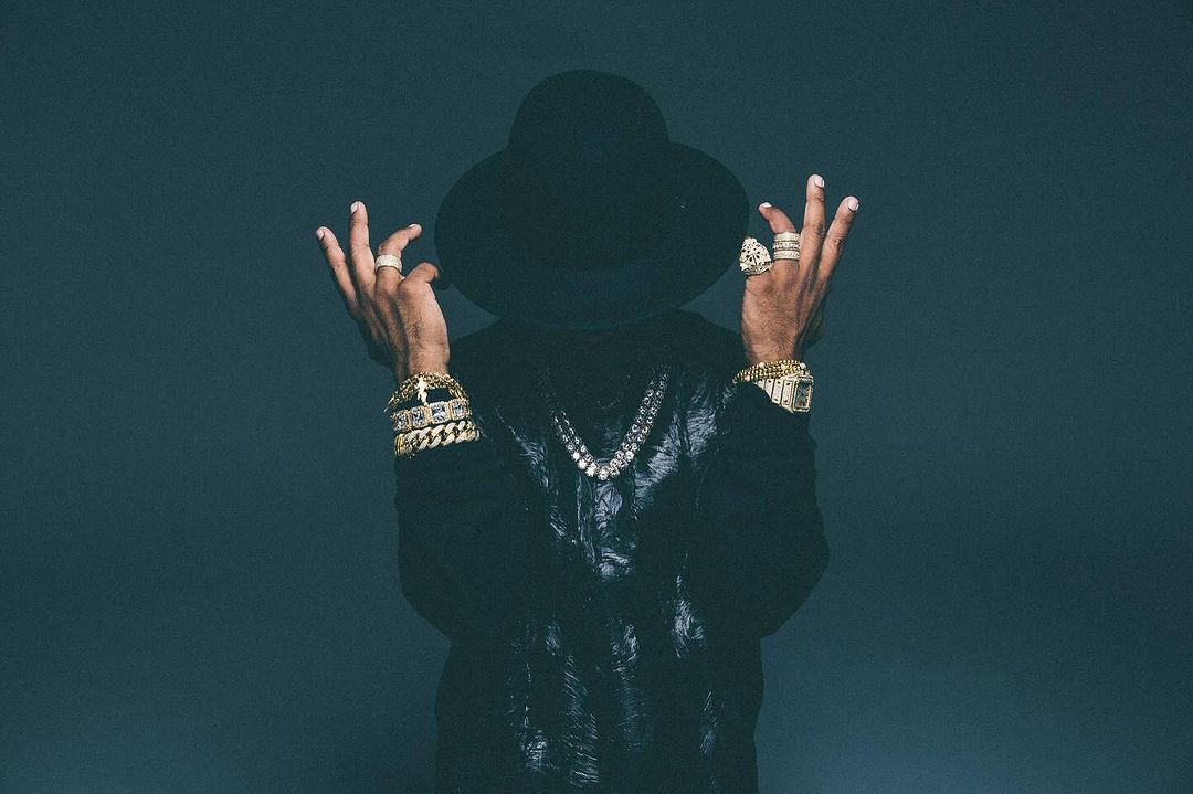 Stylish man with hat and bling poses in front of a grey backdrop, showcasing gold bracelets, rings, watch, and diamond necklace.