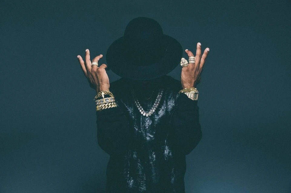 Stylish man with hat and bling poses in front of a grey backdrop, showcasing gold bracelets, rings, watch, and diamond necklace.