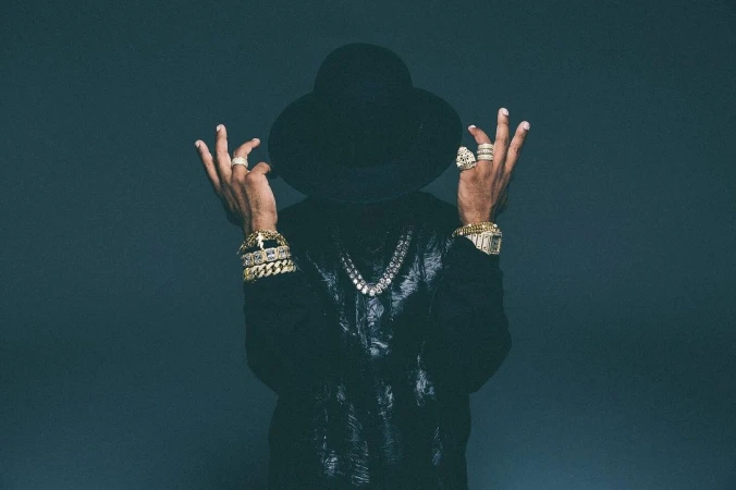 Stylish man with hat and bling poses in front of a grey backdrop, showcasing gold bracelets, rings, watch, and diamond necklace.
