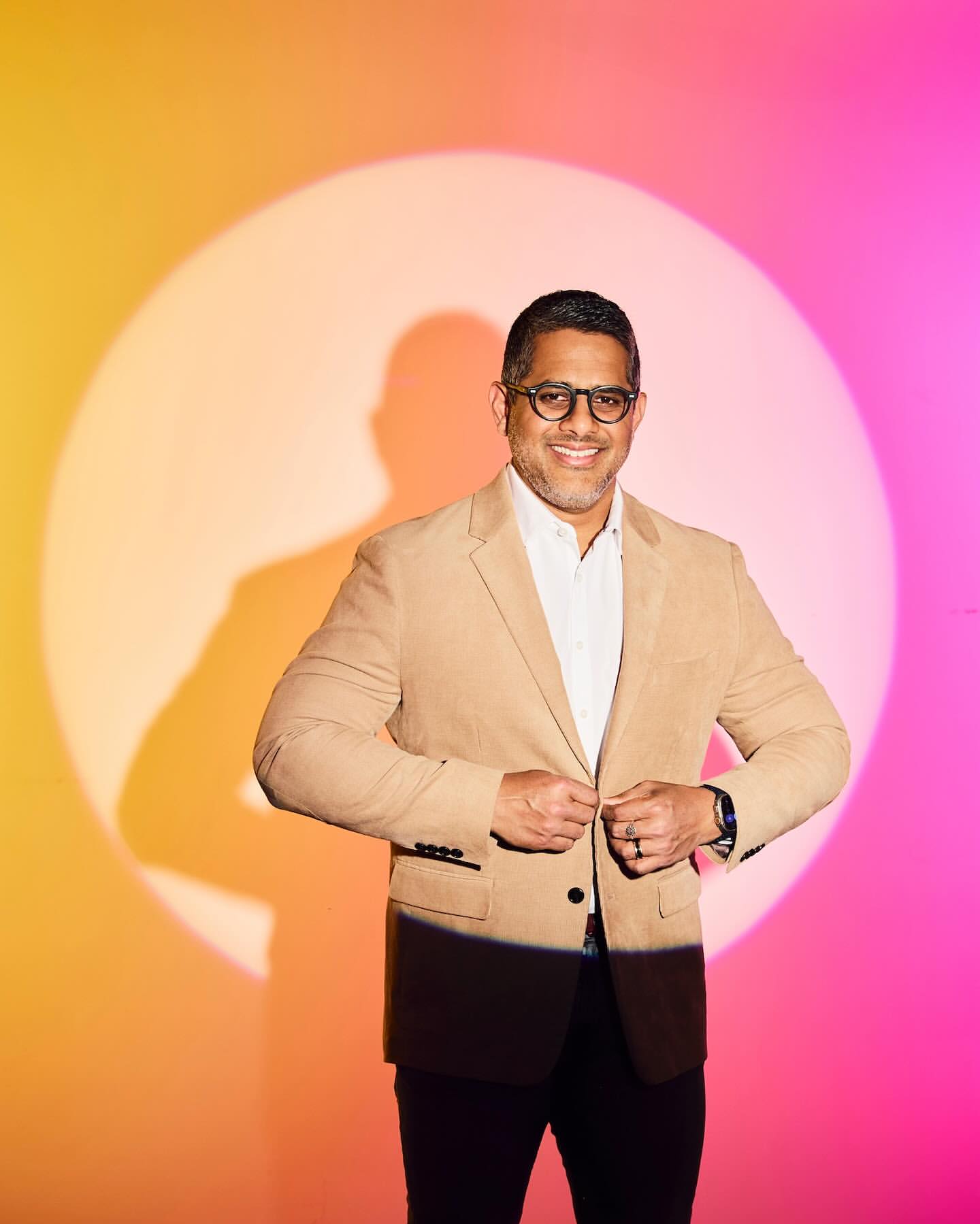 Portrait of an adult male wearing glasses and a tan blazer, buttoning the blazer, against a pink and yellow background with a light flare.