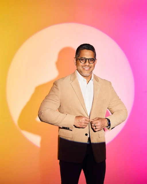 Portrait of an adult male wearing glasses and a tan blazer, buttoning the blazer, against a pink and yellow background with a light flare.